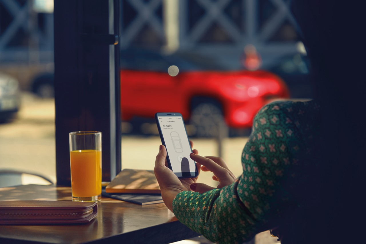 Lady sat in a cafe. She interacts with the TOYOTA MyT app on her phone, her Toyota Yaris Cross is parked in the background.